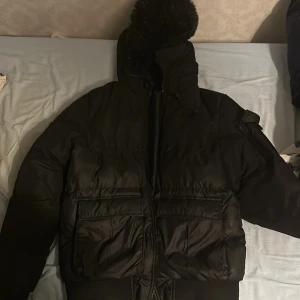 Svart pufferjacka från SikSilk - Säljer en svart pufferjacka från SikSilk i storlek S. Jackan har en mysig huva med pälsdetaljer och praktiska fickor. Den är perfekt för kalla dagar och har en stilren design med dragkedja och knappar. Passar både till vardags och festliga tillfällen.