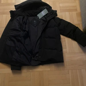 Svart pufferjacka från Canada Goose - Säljer en svart pufferjacka från Canada Goose i mycket bra skick. Jackan är perfekt för vintern med sin tjocka vaddering och långa ärmar. Den har en dragkedja och knappar framtill samt en huva för extra skydd mot kylan. Märkesloggan syns tydligt på ärmen. Perfekt för kalla dagar! ❄️