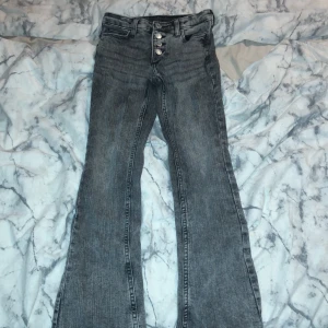 Grå bootcut jeans från H&M - Säljer ett par grå bootcut jeans från H&M i storlek EUR 134. De har låg midja och justerbar midja för perfekt passform. Jeansen är i bra skick och har en snygg knappdetalj framtill. Perfekta för en avslappnad stil!