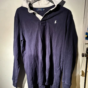 Rugbytröja från Ralph Lauren - Säljer en snygg mörkblå pikétröja från Ralph Lauren med klassisk vit krage och det ikoniska logotypbroderiet på bröstet. Perfekt för en stilren och avslappnad look. Tröjan är i bra skick och passar både till vardags och fest. Köpt för 1500kr  på Ralph Laurens hemsida. Tveka inte att höra av dig om du har frågor eller vill ha fler bilder!