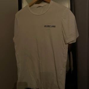 Vit Helmut Lang T-shirt - Riktigt modern vit Helmut Lang T-shirt i bra skick, i storlek S. Går att matcha till alla outfits med en elegant och tidlös logga vid bröstet som inte är för mycket. Perfekt passform och väldigt skön och mjuk att ha på sig, nypris ca 2500kr. 