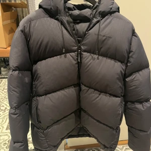Svart pufferjacka från Peak Performance - Säljer en svart pufferjacka från Peak Performance i storlek M. Jackan är perfekt för kalla vinterdagar med sin tjocka vaddering och huva. Den har en dragkedja framtill och är i mycket bra skick. Perfekt för både vardag och utomhusaktiviteter!