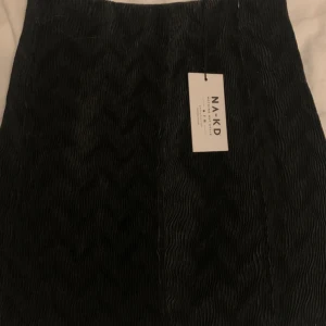 Svart kjol från NA-KD - Säljer en stilren svart kjol från NA-KD i nytt skick. Perfekt för både vardag och fest. Kjolen har en elegant struktur och är gjord i ett bekvämt material. Passar till alla säsonger och kan enkelt matchas med olika toppar och accessoarer.