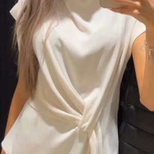 Vit blus med knytdetalj - Säljer en elegant vit blus med en snygg knytdetalj framtill. Blusen har en lös passform och är ärmlös, vilket gör den perfekt för både vardag och fest. Passar bra till både jeans och kjol för en stilren look.