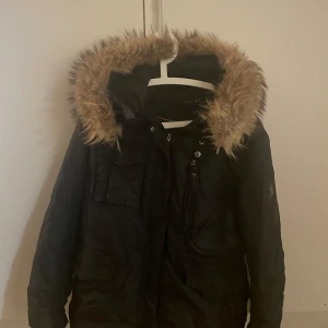 Svart parkajacka med äkta pälsdetaljer - Säljer en svart parkajacka med mysig äkta päls runt huvan. Perfekt för kalla vinterdagar. Jackan har flera praktiska fickor och stängs med dragkedja och knappar. Den är i bra skick och håller dig varm och stilren hela säsongen. Märke Hollies. 