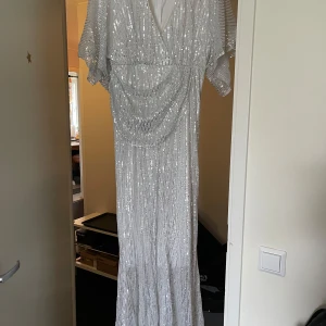 Glittrig silverklänning - Säljer en fantastisk glittrig silver-vit  jumpsuit med v-ringning och kortärmade ärmar. Perfekt för nyårsfesten eller en glamorös kväll.Jumpsuit  har en elegant drapering framtill och är aldrig använd . Den är lång och ger en riktigt lyxig känsla. Passar perfekt för dig som vill glänsa!