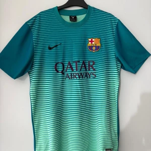 Turkos Barcaelona t shirt - Säljer en snygg turkos fotbollströja från Nike med FC Barcelona-emblem och Qatar Airways-logga på framsidan. Tröjan har korta ärmar och ett randigt mönster i ljusare turkos nyans. Perfekt för fotbollsfans som vill visa sitt stöd! 🏆