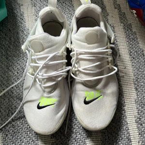 Vita Nike sneakers - Säljer ett par vita Nike sneakers i bra skick. De har en stilren design med svarta och neongula detaljer. Skorna har snörning och är perfekta för både vardag och träning. De är bekväma och lätta, vilket gör dem idealiska för en aktiv livsstil.