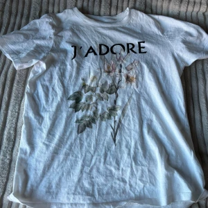 Vit t-shirt med blommotiv från Gina Tricot - Säljer en superfin vit t-shirt från Gina Tricot i storlek XS. Den har ett tryck med texten 'J'ADORE' och ett vackert blommotiv i mjuka färger. Perfekt för en avslappnad look eller att styla upp med en snygg jacka. T-shirten är i bra skick och gjord av mjukt bomullsmaterial.