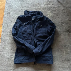 Mörkblå jacka från Indicode - Säljer en snygg mörkblå jacka från Indicode i storlek medium. Jackan har en stilren design med dragkedja och knappar framtill. Perfekt för höst och vår med sina praktiska fickor och bekväma passform. Passar både till vardags och lite finare tillfällen. 🧥