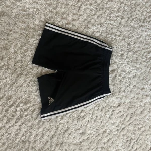 Svarta shorts från Adidas - Säljer ett par svarta Adidas shorts i storlek 164. De har de klassiska tre vita ränderna längs sidorna och är perfekta för träning eller fotboll. Materialet är lätt och andas bra, vilket gör dem bekväma att bära under varma dagar. Passar perfekt för sommarens aktiviteter! Helt nya säljer pga fel storlek för mig 