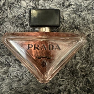 Parfym från Prada (Milano) (oanvänd) 90ml - Säljer en lyxig parfym från Prada i en unik trekantig flaska. Flaskan är genomskinlig med en svart kork och har en elegant design. Perfekt för den som vill ha en exklusiv doftupplevelse. Passar både till vardags och fest!