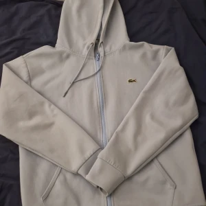 Ljus blå hoodie från Lacoste - Säljer en stilren Ljusblå hoodie från Lacoste med dragkedja och klassisk krokodillogga på bröstet. Tröjan har en huva med dragsko och långa ärmar. Perfekt för en avslappnad look. Bra skick sista pris 800