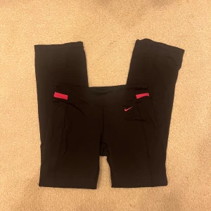 Flare träningsbyxor från Nike - Snygga svarta träningsbyxor från Nike med en röd detalj vid midjan. Byxorna har en tight passform och är perfekta för träning. Tillverkade i ett bekvämt syntetmaterial med Dri-FIT-teknologi för att hålla dig torr. Använd endast en gång, står M på etiketten men sitter som S
