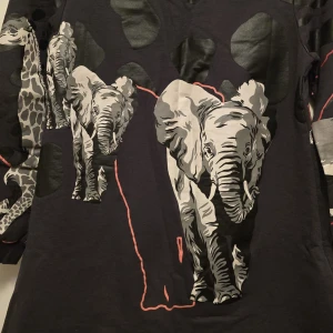 Svart tröja med djurmotiv från Kenzo - Säljer en svart tröja från Kenzo med unika djurmotiv av elefanter och tigrar. Tröjan har långa ärmar och ett coolt mönster med röda detaljer. Perfekt för dig som vill sticka ut med en djärv stil!