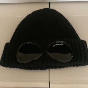 Svart ribbad mössa från C.P. Company - Snygg svart ribbad mössa från C.P. Company med unika glasögondetaljer framtill. Perfekt för att hålla värmen med stil. Mössan har en klassisk design med uppvikt kant.