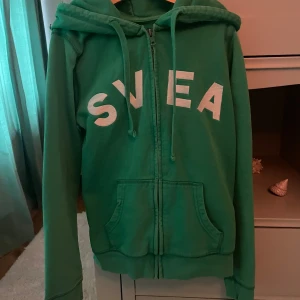 Grön hoodie från Svea - Säljer en grön hoodie från Svea med dragkedja och stor logga på framsidan. Den har en justerbar huva med snören och praktiska fickor framtill. Perfekt för en avslappnad stil. Tröjan är i storlek s men jag skulle säga att den passar både XS och XXS
