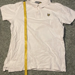 Vit pikétröja från Lyle & Scott - Säljer en klassisk vit pikétröja från Lyle & Scott med korta ärmar och en broderad logga på bröstet. Tröjan har en stilren design med krage och knappar framtill. Perfekt för en avslappnad och stilren look.Tröjan passar även bra på 13 åringar 