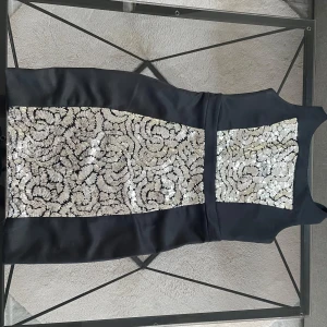 Svart klänning med paljetter från SisterS Point - Snygg svart klänning från SisterS Point med glittrande silverpaljetter framtill. Klänningen har en tight passform och är ärmlös, perfekt för en festlig kväll. Den är knälång och har en elegant design som verkligen sticker ut.