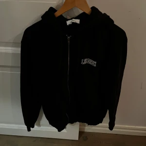 Svart hoodie från Les Deux - Säljer en stilren svart hoodie från Les Deux med dragkedja framtill. Den har en liten logotyp på bröstet och en bekväm huva. Perfekt för en avslappnad stil. Beövs strykas.