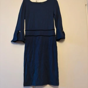 Mörkblå klänning med dekorativa knappar - Elegant mörkblå klänning med långa ärmar och dekorativa knappar vid ärmsluten. Klänningen har en stilren design med en markerad midja och en klassisk siluett. Perfekt för en stilfull look.