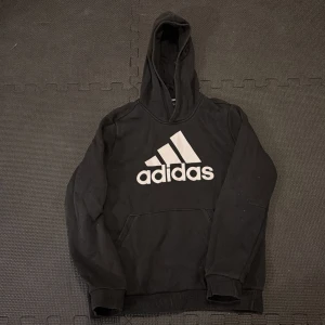 Svart hoodie från Adidas - Säljer en klassisk svart hoodie från Adidas med det ikoniska logotyptrycket på framsidan. Tröjan har en stor huva och en praktisk magficka. Lite blekt. Liten i strl. Ca 140 ist. 