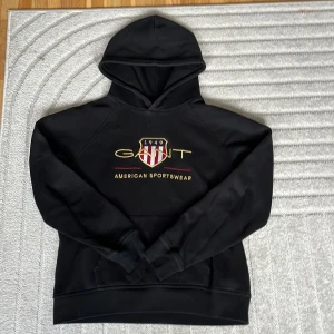 GANT Svart hoodie - Säljer en svart hoodie från Gant med deras klassiska logga och texten 'American Sportswear' på framsidan. Tröjan har en bekväm huva och långa ärmar, perfekt för en avslappnad stil. storlek varierar beroende på passform. Pris kan diskuteras
