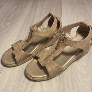 Beige sandaler med kilklack från Michael Kors - Snygga beige sandaler från Michael Kors med kilklack och dragkedja framtill. Sandalerna har ett diskret mönster och en bekväm passform. Perfekta för sommarens alla tillfällen. Pris får diskuteras!