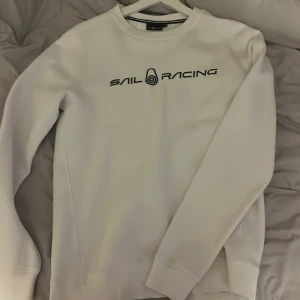 Vit tröja från Sail Racing - Säljer en stilren vit tröja från Sail Racing med deras logga tryckt på framsidan. Tröjan har långa ärmar och en klassisk rund halsringning. Perfekt för en avslappnad och sportig look.