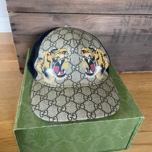 Gucci keps - Snygg keps från Gucci med ett tigertryck och det ikoniska GG-mönstret. Kepsen har en beige bas med svarta mesh-detaljer på sidorna för extra ventilation. Perfekt för att ge din outfit en lyxig touch. Självklart äkta.