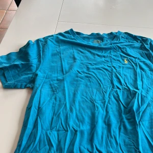 Blå t-shirt från Ralph Lauren - Säljer en blå t-shirt från Ralph Lauren med det klassiska broderade logotypen i gult på bröstet. T-shirten har en normal passform och är kortärmad, perfekt för en avslappnad stil.