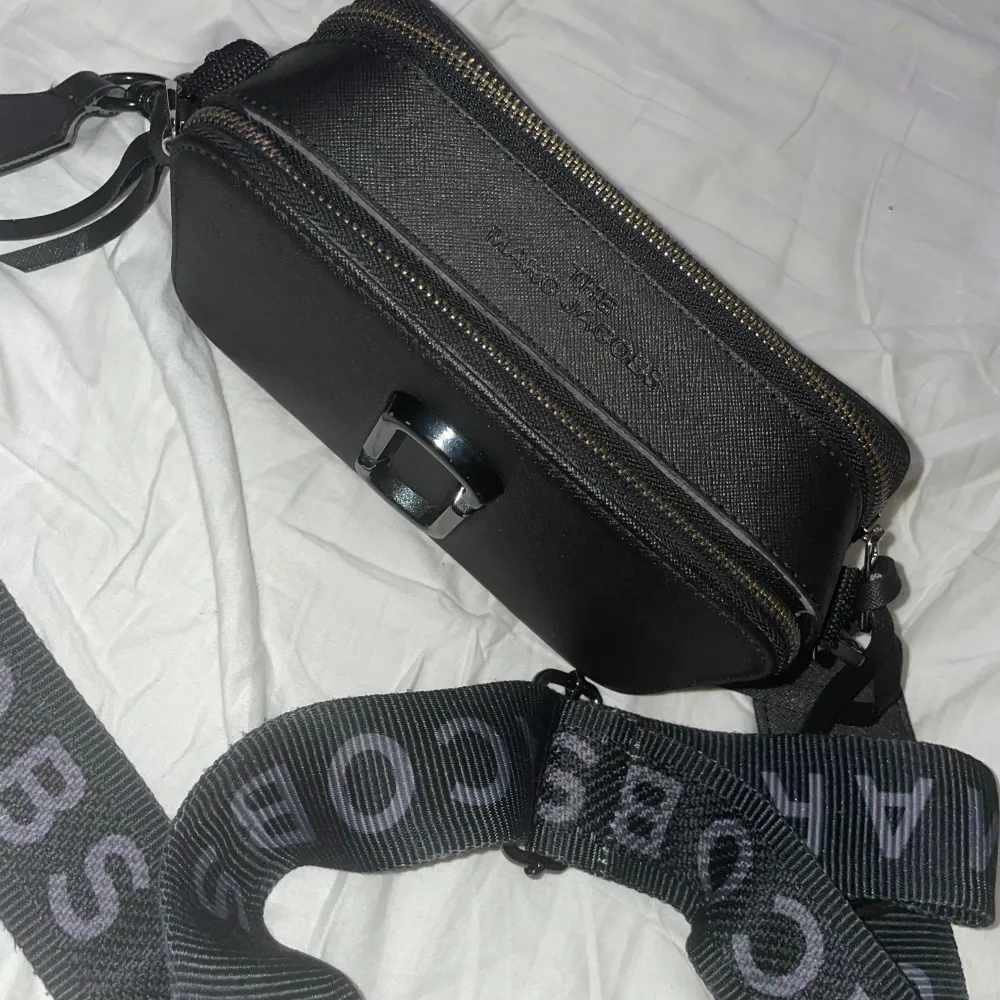 Snygg svart axelväska från Marc Jacobs med ett stilrent design. Väskan har en justerbar axelrem med märkets logga och två dragkedjefack för enkel organisering. Perfekt för att bära dina nödvändigheter med stil.. Laukut & Käsilaukut.
