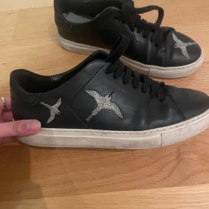 Svarta Axel Arigato sneakers - Snygga Axel arigato sneakers med vita sulor och ett fågelmotiv i silver på sidan. Skorna har snörning och är som ny!