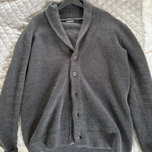 Grå kofta från John Henric - Säljer en stilren grå kofta från John Henric i storlek M. Koftan har en ribbad struktur och knappar framtill. Perfekt för kyliga dagar och ger en klassisk look.