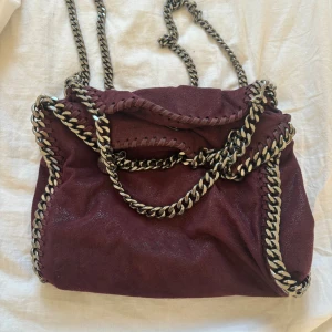 Vinröd axelväska med kedjedetaljer - Snygg vinröd axelväska från Stella McCartney med coola kedjedetaljer. Väskan har en magnetknapp för enkel stängning och en stilren design. Perfekt för att ge din outfit en edgy touch.