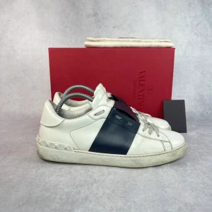 Valentino sneakers i vitt och blått - Snygga Valentino sneakers i vitt med en bred blå rand. Skorna har klassisk snörning och dekorativa nitar runt hälen. Perfekta för en stilren look.