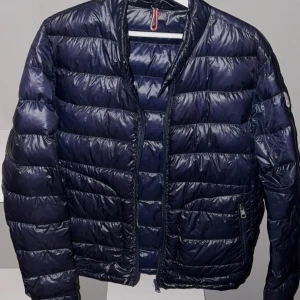 Mörkblå dunjacka från Moncler - Snygg mörkblå dunjacka från Moncler med dragkedja framtill. Jackan har en quiltad design och långa ärmar, perfekt för kyligare dagar. Den är stilren och passar bra till olika outfits. 🧥