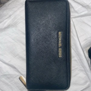 Svart plånbok från Michael Kors - Snygg svart plånbok från Michael Kors med dragkedja runtom. Tillverkad i skinn med guldfärgade detaljer. Perfekt för att hålla ordning på kort och kontanter. En stilren accessoar för alla tillfällen.