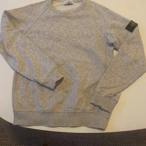 Grå sweatshirt från Stone Island - Säljer en grå sweatshirt från Stone Island med deras ikoniska märke på ärmen. Tröjan har långa ärmar och ribbade muddar. Perfekt för en avslappnad stil. Hör av er vid funderingar!