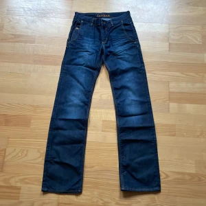 Mörkblå vintage jeans från East West - (W26/L33) Snygga mörkblå jeans från East West med klassisk femficksdesign och knappdetaljer på bakfickorna. Jeansen har en rak passform och är perfekta för en avslappnad stil. De har en dragkedja och knapp framtill. 