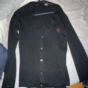 Svart kofta från Alexander McQueen - Snygg svart kofta från Alexander McQueen med knappar framtill och två fickor. Den har en broderad logga i rött på bröstet och är stickad för en bekväm känsla. Perfekt för att lägga till en touch av stil till din outfit.