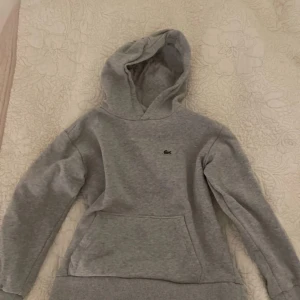 Grå hoodie från Lacoste - Säljer en klassisk grå hoodie från Lacoste med den ikoniska krokodilloggan på bröstet. Tröjan har en bekväm huva och en stor ficka framtill. Perfekt för en avslappnad stil. 🐊