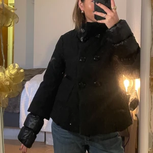 Svart dunjacka från Benetton - Snygg svart dunjacka från Benetton med dubbelknäppning och hög krage. Jackan är vadderad med 80% dun och 20% fjäder för extra värme. Perfekt för kalla dagar med sin stilrena design och långa ärmar.