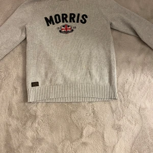 Grå tröja från Morris - Säljer en stilren grå tröja från Morris med svart text och en broderad brittisk flagga på framsidan. Tröjan har långa ärmar och ribbade kanter vid ärmslut och nederkant. Perfekt för en avslappnad och snygg look.