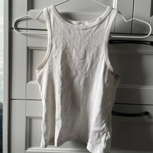 Vit ribbad linnetopp - Säljer en stilren vit ribbad linnetopp. Den är ärmlös och har en klassisk rund halsringning. Perfekt för varma dagar eller som lager under en tröja. Tyvärr har den blivit lite gulaktig under armarna. Storlek 152.