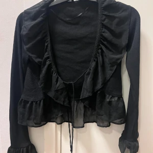 Svart ribbad kofta med volangdetaljer - Snygg svart kofta med ribbad textur och volangdetaljer. Den har långa ärmar och knytband framtill. Perfekt för att lägga till en touch av stil till din outfit. 💕