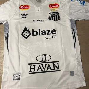 Vit fotbollströja från Umbro - Snygg vit fotbollströja från Umbro med tryck av olika sponsorer och klubbmärke på framsidan. Tröjan har korta ärmar och nummer 10 med ett namn på baksidan. Perfekt för fotbollsfans som vill visa sitt stöd.