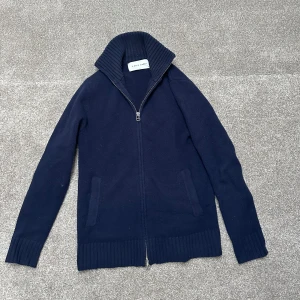 Mörkblå kofta från A Day's March - Säljer en stilren mörkblå kofta från A Day's March i storlek S. Koftan har en dragkedja framtill och ribbade detaljer vid krage, ärmslut och nederkant. Perfekt för kyliga dagar och enkel att matcha med olika outfits.