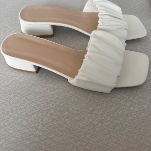 Vita sandaler med klack - Stilrena vita sandaler med liten klack. De har en bred, rynkad rem över foten som ger en modern touch. Perfekta för sommarens alla tillfällen. Helt oanvända! Storlek 39