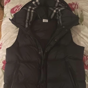 Svart dunväst från Burberry - Snygg svart dunväst från Burberry med dragkedja och knappar. Västen har en klassisk Burberry-rutig insida på huvan och är perfekt för kyligare dagar. Den har en stilren design och är både praktisk och trendig. Går att göra till jacka. ÄKTA 